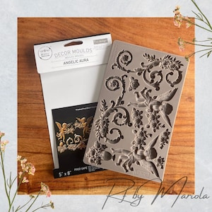 May include: A silicone mould for baking or crafting, featuring a design of angels and floral scrolls. The mould is grey and has the text "Angelic Aura" printed on it. The mould is 5 inches by 8 inches and is food safe.