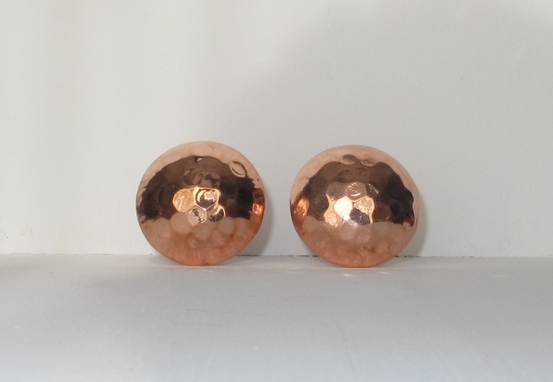May include: A pair of copper earrings with a hammered finish. The earrings are round and have a slightly domed shape.