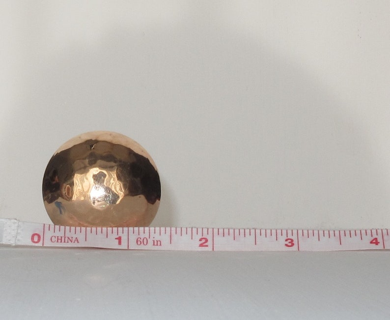 May include: A hammered copper bowl with a shiny finish. The bowl is about 1 inch in diameter and is sitting on a white surface next to a measuring tape.