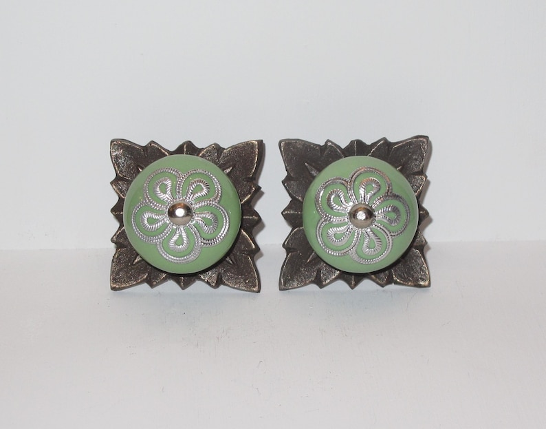 Peut inclure: Deux boutons de cabinet en c&eacute;ramique verte avec des motifs floraux argent&eacute;s, mont&eacute;s sur des bases en m&eacute;tal bronze orn&eacute;es.