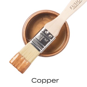 May include: A small jar of Fusion Mineral Paint in the color Copper. A paintbrush is resting on top of the jar, with the bristles dipped in the paint.