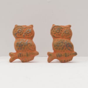 May include: A pair of orange owl figurines with silver detailing. The owls are perched on a branch and have their wings spread out.