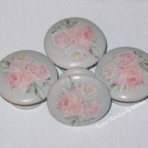 Pode incluir: Quatro botões de madeira pintados de cinza com desenhos florais rosa e branco. Os botões são redondos e têm um acabamento liso. "Renewal Furniture by Mariola" está escrito em preto no botão inferior direito.