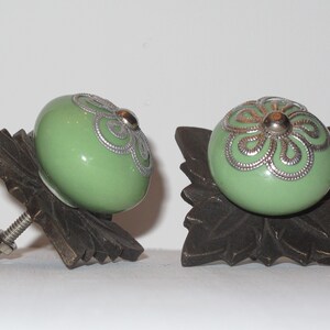 Peut inclure: Deux boutons de cabinet en c&eacute;ramique verte avec des motifs floraux argent&eacute;s, mont&eacute;s sur des bases en bronze en forme de feuille orn&eacute;es.