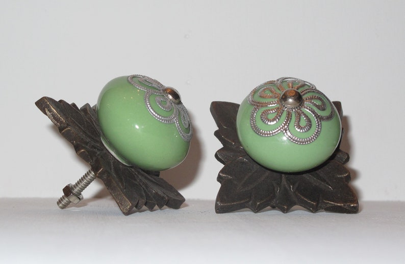 Peut inclure: Deux boutons de cabinet en c&eacute;ramique verte avec des motifs floraux argent&eacute;s, mont&eacute;s sur des bases en bronze en forme de feuille orn&eacute;es.