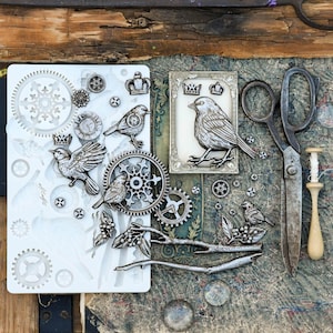 May include: A white silicone mould featuring various raised designs including birds, gears, crowns, branches, and leaves. Beside the mould are several metallic-coloured cast pieces of these shapes, a rectangular frame, vintage metal scissors, and a wooden tool.