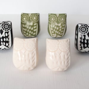May include: Six ceramic owl figurines in various colours, including black, white, and green. Each owl has a unique design with white and black details. The owls are all facing forward and have their wings spread out.