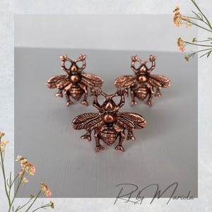 以下が含まれることがあります： ブロンズ色のハチの形をしたキャビネットノブが3つ。ノブには羽、脚、触角が詳細に描かれています。ノブは薄い灰色の表面に配置されています。これらの装飾的なノブは、家具にユニークなタッチを加えます。