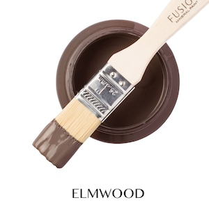 May include: A small jar of Fusion Mineral Paint in the color Elmwood. The jar is open and a paintbrush is resting on the rim of the jar. The brush is partially dipped in the paint.