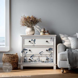 May include: A white three-drawer chest with black knobs and bird and marsh-themed decals. The chest is decorated with blue and white illustrations of birds, trees, and marsh plants. A woven basket and a vase with dried flowers are on top of the chest. The text at the bottom reads "#682804 whispers by the marsh."