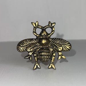 Może przedstawiać: Złota metalowa figurka pszczoły z misternymi detalami. Pszczoła ma fakturowane ciało i skrzydła, a jej nogi są wyciągnięte.