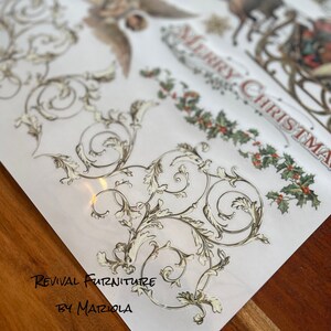 Op de afbeelding: Een vel decoratief papier met een gouden en wit bloemmotief. Het papier heeft de tekst "Revival Furniture by Mariola" erop gedrukt. Het papier ligt op een houten oppervlak.