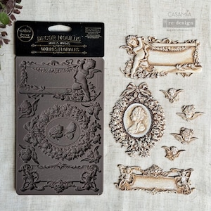 May include: A selection of decorative moulds and castings. The dark grey silicone mould features ornate designs, including cherubs, frames, and floral elements. Several finished castings are displayed, showcasing the intricate details and textures.