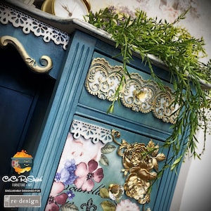 May include: A teal blue dresser with gold accents and floral details. The dresser has a white lace trim and a floral design on the front. The dresser is decorated with gold roses and crystal knobs.