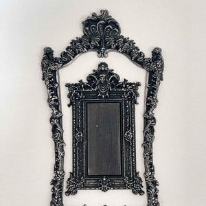 Op de afbeelding: Een zwarte, sierlijke fotolijst met een kleinere zwarte, sierlijke fotolijst erin. Beide lijsten hebben een decoratief ontwerp met bloemmotieven.