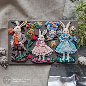 May include: A grey silicone mould with Easter designs. It features detailed bunny figures in Victorian clothing, Easter eggs, and floral accents. The text "#681692 Decor Moulds Meadow Whispers" is visible.