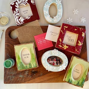 May include: An assortment of decorated cookies, some shaped like frames, with messages like "Thank You" and "Merry Christmas." The cookies are on a wooden board, with a brush, glitter, and small cards.