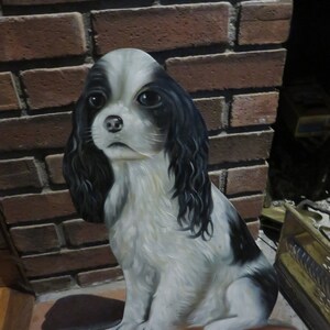 May include: A black and white painted figurine of a dog sitting on a brown and tan cushion. The dog has black fur and white markings on its face and chest.