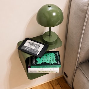 May include: A wall-mounted olive green metal floating shelf holding a matching olive green dome-shaped table lamp, a black e-reader, and two books titled In a Dark Dark Wood by Ruth Ware and a book with the text EMOЦIЙНИЙ IHTEЛEKT on the spine.