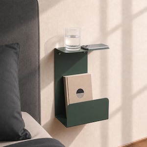 May include: A dark green metal wall-mounted bedside shelf featuring a flat top surface for a glass of water and a smartphone, and a lower pocket compartment for storing a book. The shelf is attached to a wall next to a dark gray upholstered bed.