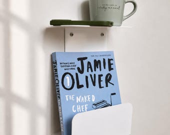 May include: A white metal wall-mounted shelf with two tiers. The top tier holds a light green mug with the text "see the good in every moment" and a dark green phone case. The bottom tier holds a blue book titled "Jamie Oliver The Naked Chef".