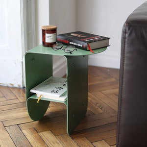 Op de afbeelding: Een groene metalen bijzettafel met een afgerond, open ontwerp. De tafel heeft twee planken en heeft een boek, een kaars en een bril. De tafel staat naast een bruine leren bank.