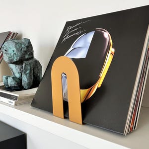 May include: A shelf displays vinyl records, a sculpture, and decorative bookends. The bookends are in the shape of a U, one in a muted gold color and the other in a muted green color. The album cover on the right features a helmet design.