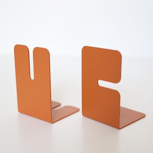 Op de afbeelding: Een paar L-vormige boekensteunen van metaal met een matte oranje afwerking. Elke boekensteun heeft een opvallend uitgesneden ontwerp, waarbij de ene een verticale gleuf heeft en de andere een horizontale inkeping, wat een stabiele basis biedt.