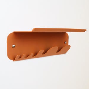Könnte beinhalten: Wandregal aus gebranntem orangefarbenem Metall mit eingebautem Regal und einer Reihe von wellenförmigen Haken. Das Regal ist mit zwei silbernen Schrauben an einer weißen Wand befestigt. Minimalistisches und modernes Design.