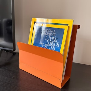Könnte beinhalten: Ein orangefarbener Metall-Zeitschriftenhalter mit rechteckiger Form. Er enthält mehrere Zeitschriften, darunter eine mit blauem Cover und dem Text "NATIONAL GEOGRAPHIC LIVING LONGER AND BETTER". Der Halter steht auf einer dunklen Oberfläche.