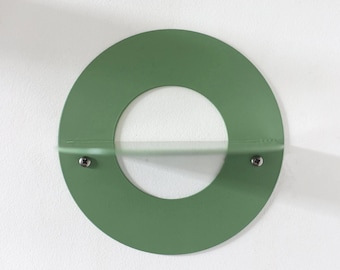 May include: A circular sage green metal wall shelf featuring a central horizontal ledge. The design includes a hollow middle section and two visible silver-toned screws securing the shelf to the wall surface for mounting purposes.