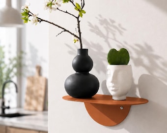 Könnte beinhalten: Ein wandmontiertes, terrakottafarbenes Regal mit einer halbkreisförmigen Halterung. Auf dem Regal stehen eine mattschwarze, kürbisförmige Vase mit einem blühenden Zweig und ein weißer Keramikübertopf in Form eines Gesichts mit einem herzförmigen Blatt.