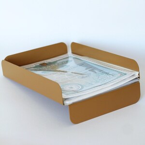 May include: A matte gold metal document tray with rounded corners and low side walls, containing a stack of white papers with blue printed text and a circular seal, designed for organising office documents on a flat surface.