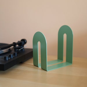 May include: A sage green metal bookend with a rounded arch design. The bookend is on a wooden surface, with a black record player in the background. The bookend is approximately 6 inches wide and 6 inches tall.