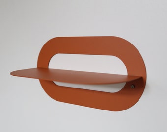 May include: A wall-mounted shelf featuring a burnt orange metal frame with a horizontal oval cutout. A flat, matching orange shelf surface extends outward from the center of the oval frame, secured by a small visible screw on the right side.