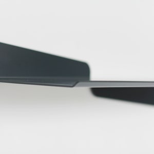 May include: A minimalist, dark gray metal shelf mounted on a white wall. The shelf has a simple, modern design with a flat surface and two angled supports. The shelf is designed for displaying items.