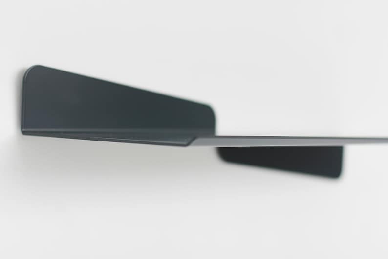 May include: A minimalist, dark gray metal shelf mounted on a white wall. The shelf has a simple, modern design with a flat surface and two angled supports. The shelf is designed for displaying items.