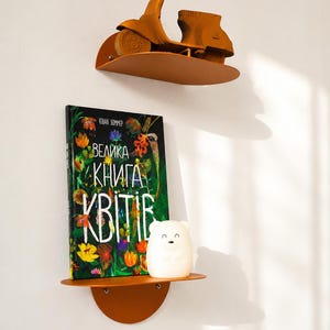 May include: Two semi-circular orange metal wall shelves. The top shelf holds a brown scooter model. The bottom shelf holds a book titled "ВЕЛИКА КНИГА КВІТІВ" with a colorful floral cover and a small white bear-shaped night light.