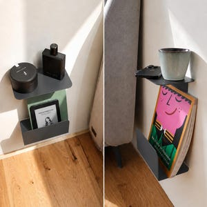 Op de afbeelding: Een donkergrijze metalen wandplank met een plat bovenoppervlak en een onderste vak. De plank bevat diverse voorwerpen, waaronder een zwarte container, een zwarte fles, een e-reader, een smartphone, een keramische beker en een boek.