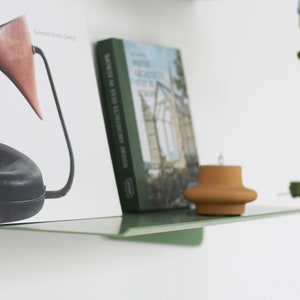 May include: A modern, minimalist green shelf with books and a small terracotta-colored candle holder. One book is titled "Selected Nordic Design," and the other is titled "Where Architects Stay in Europe." A trailing green plant is visible.