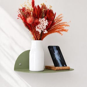 Puede incluir: Una estantería de pared semicircular de color verde salvia que sostiene un jarrón de cerámica blanco lleno de un ramo de flores secas rojas, naranjas y crema, junto a un smartphone negro sobre un pequeño soporte de madera que muestra las 15:21.