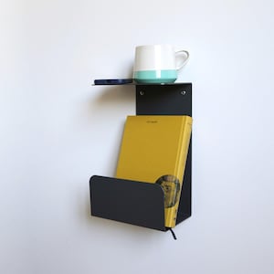 Peut inclure: Une étagère murale en métal noir avec une petite étagère supérieure contenant un téléphone bleu et une tasse blanche et turquoise. L'étagère inférieure contient un livre jaune. L'étagère est fixée à un mur blanc.