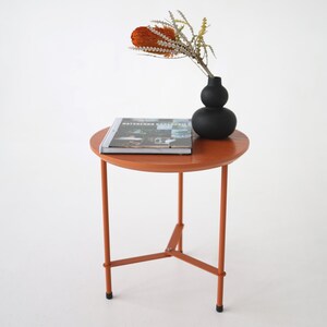 Puede incluir: Una pequeña mesa auxiliar redonda naranja con tres patas. Un jarrón negro con un arreglo floral seco y una revista encima. La mesa es de metal y tiene un diseño sencillo y moderno.