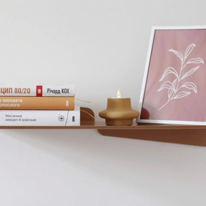 May include: A modern, minimalist shelf in a warm brown hue, holding a stack of books with titles in a foreign language, a framed botanical print, and a gold-colored candle. The shelf is mounted on a white wall.