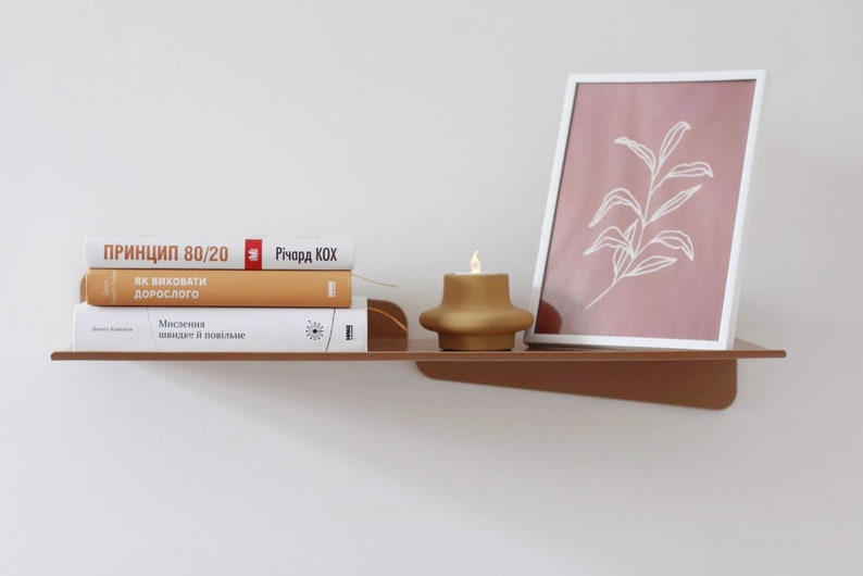 May include: A modern, minimalist shelf in a warm brown hue, holding a stack of books with titles in a foreign language, a framed botanical print, and a gold-colored candle. The shelf is mounted on a white wall.