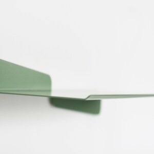 May include: A minimalist, wall-mounted shelf in a muted green hue. The shelf is made of a single piece of metal, with a flat surface and a raised edge. The shelf is approximately 12 inches long and 6 inches wide, ideal for displaying small items.