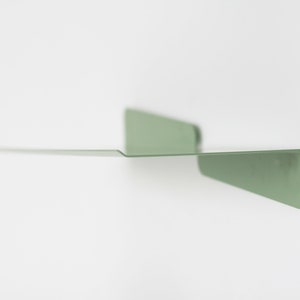 May include: A minimalist, sage green metal wall shelf. The shelf has a long, flat surface extending from two triangular supports. The design is simple and modern, suitable for displaying small items or decor. The shelf is mounted on a white wall.