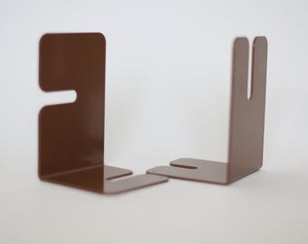 May include: Two brown metal bookends. Each bookend has a rectangular shape with a cut-out design. The bookends are positioned on a white surface, showcasing their minimalist and modern aesthetic.