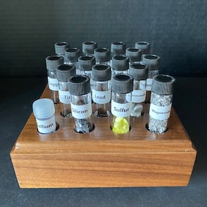 May include: A wooden display case with 20 small glass vials containing various elements. The vials are labeled with the element names, including Titanium, Silicon, Lead, Sulfur, and Magnesium.