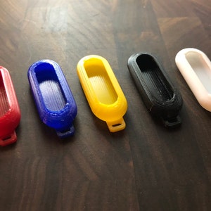 May include: Five 3D printed keychains in different colors: red, blue, yellow, black, and white. Each keychain has a smooth, rounded shape with a small loop at the top.